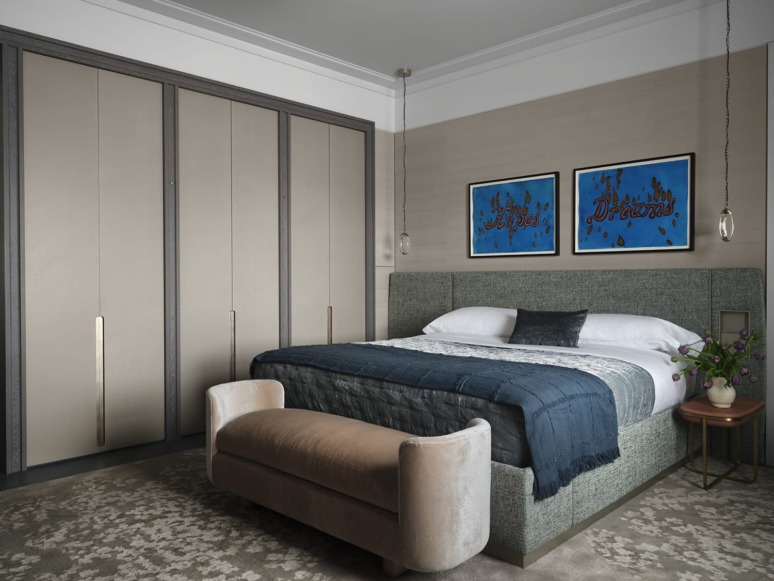 Modern bedroom with gray upholstered bed, blue bedding, large closet, artwork on walls, and a soft bench at the foot of the bed.