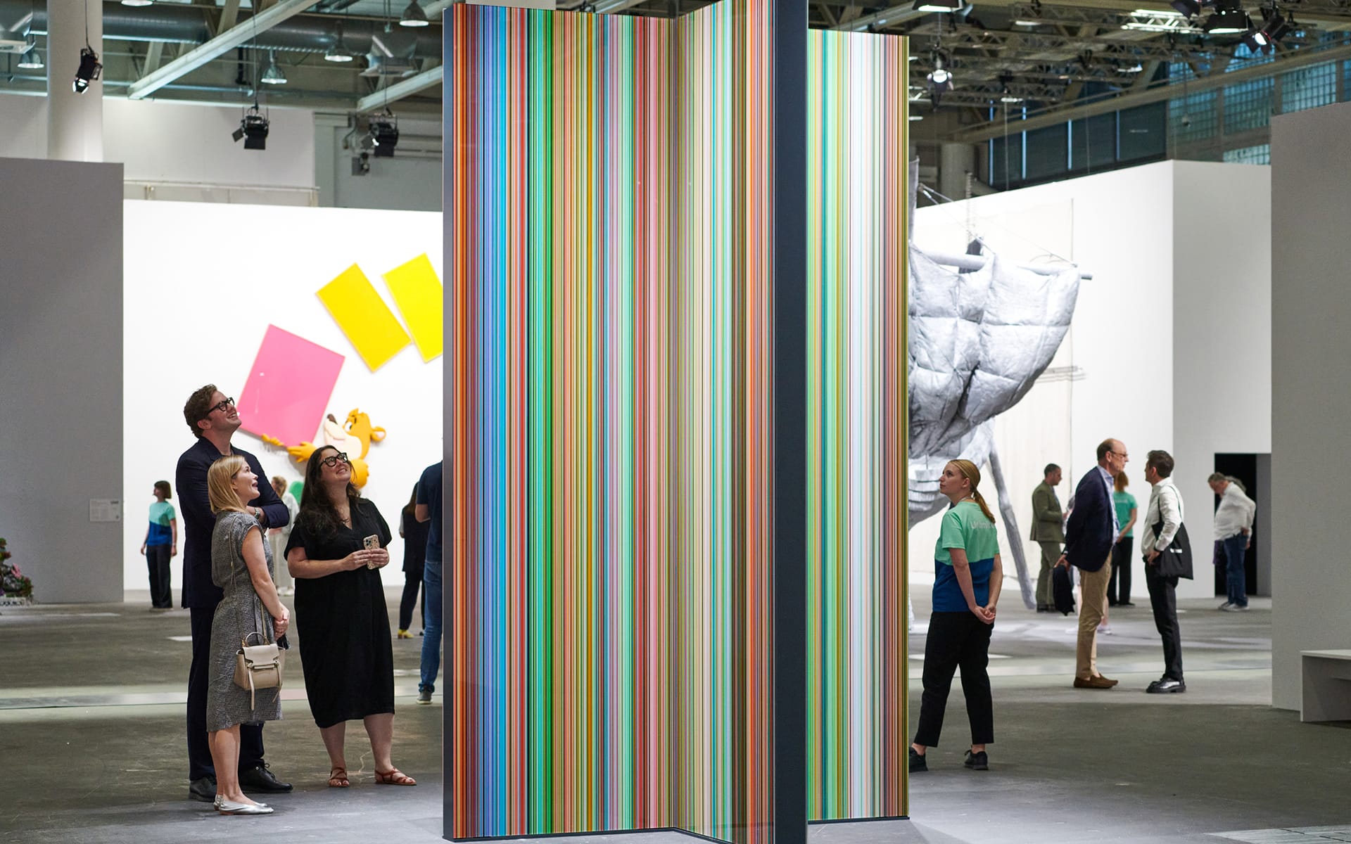 Visitors in an art gallery observe colorful vertical striped installation with abstract art and sculptures in the background.