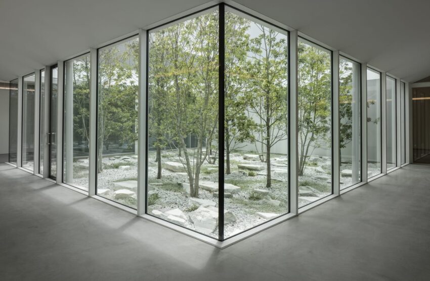 Modern building interior with glass walls surrounding a minimalist courtyard garden featuring trees and white stones.