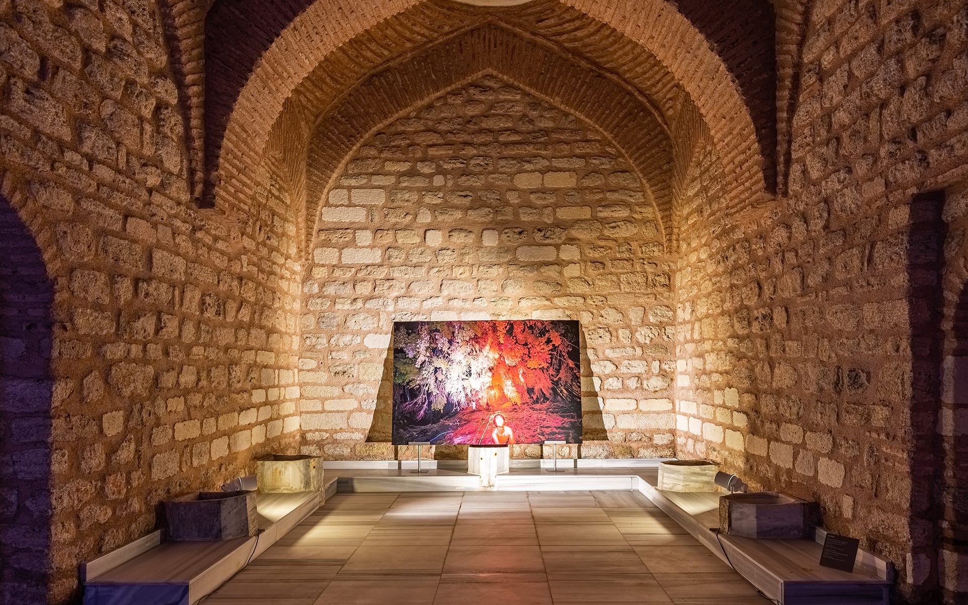 Person standing in a historic stone archway, illuminated by colorful projection art on a tapestry hanging on the back wall.
