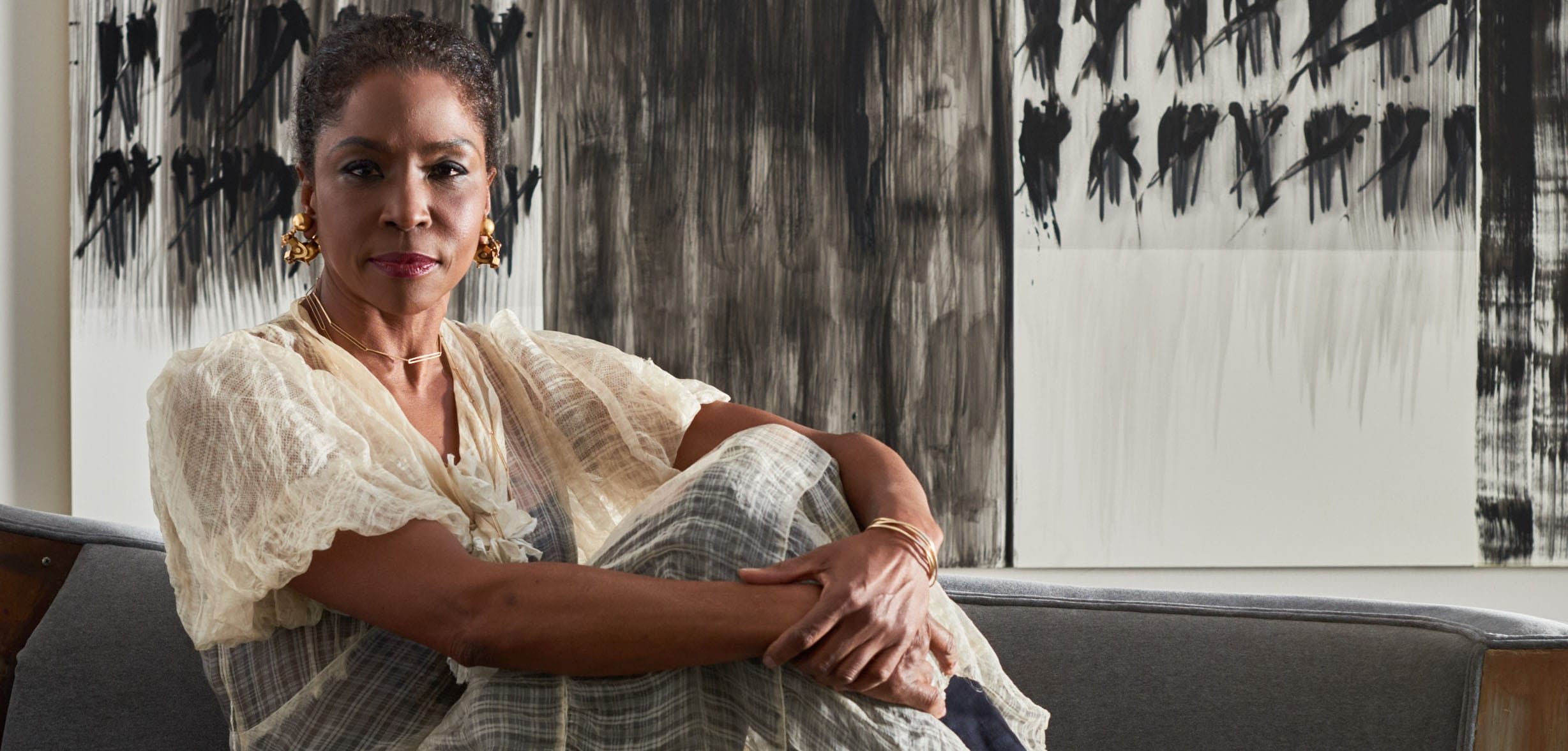 Person sitting on a couch in front of abstract black and white artwork, wearing a light-colored blouse and gold earrings.