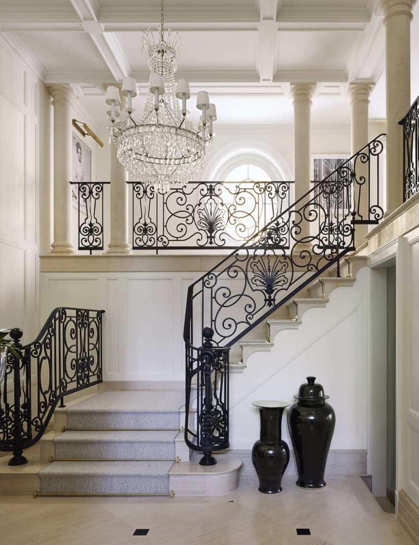 Elegant foyer with intricate wrought iron staircase and chandelier, featuring pillars and decorative vases.