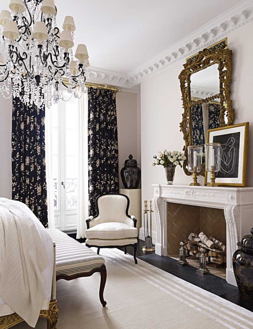 Elegant bedroom with a chandelier, mirror above fireplace, patterned curtains, and a cozy armchair near a large window.