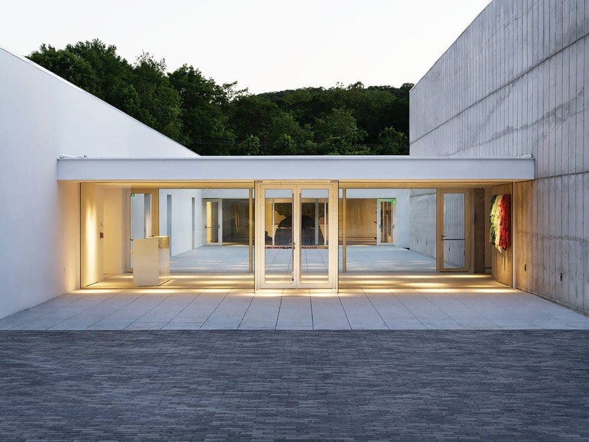 Modern building entrance with large glass doors, surrounded by trees and concrete walls, illuminated by soft lighting.