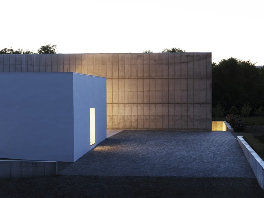 Modern minimalist building exterior at dusk with illuminated entrance, concrete and white walls, surrounded by trees.