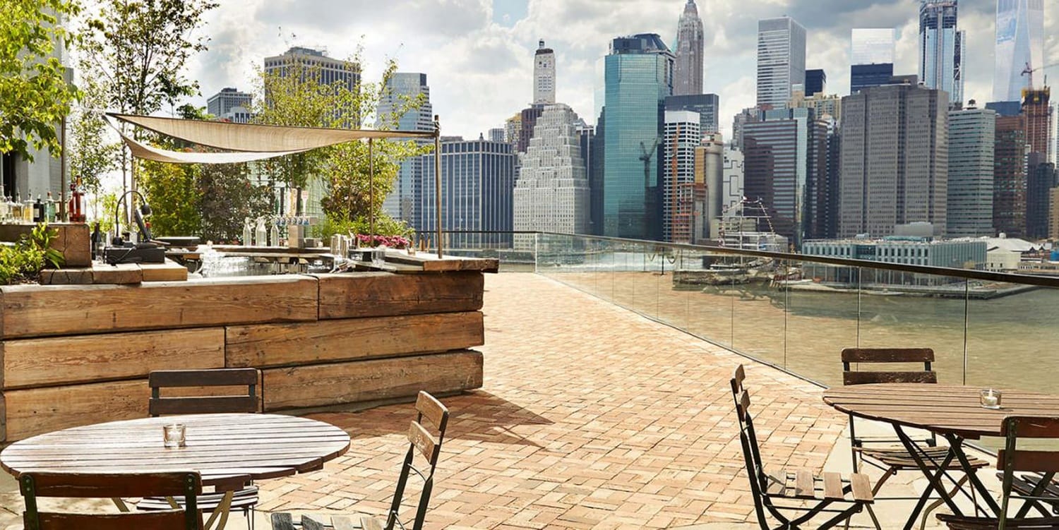 Rooftop patio with wooden tables and chairs overlooking a city skyline with tall buildings and cloudy sky.