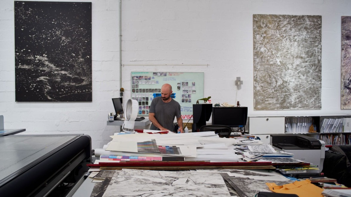 Person standing in an art studio surrounded by large canvases, papers, and office equipment.