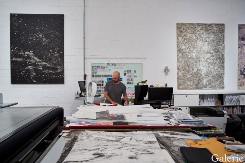 Person standing in an art studio surrounded by large canvases, papers, and office equipment.