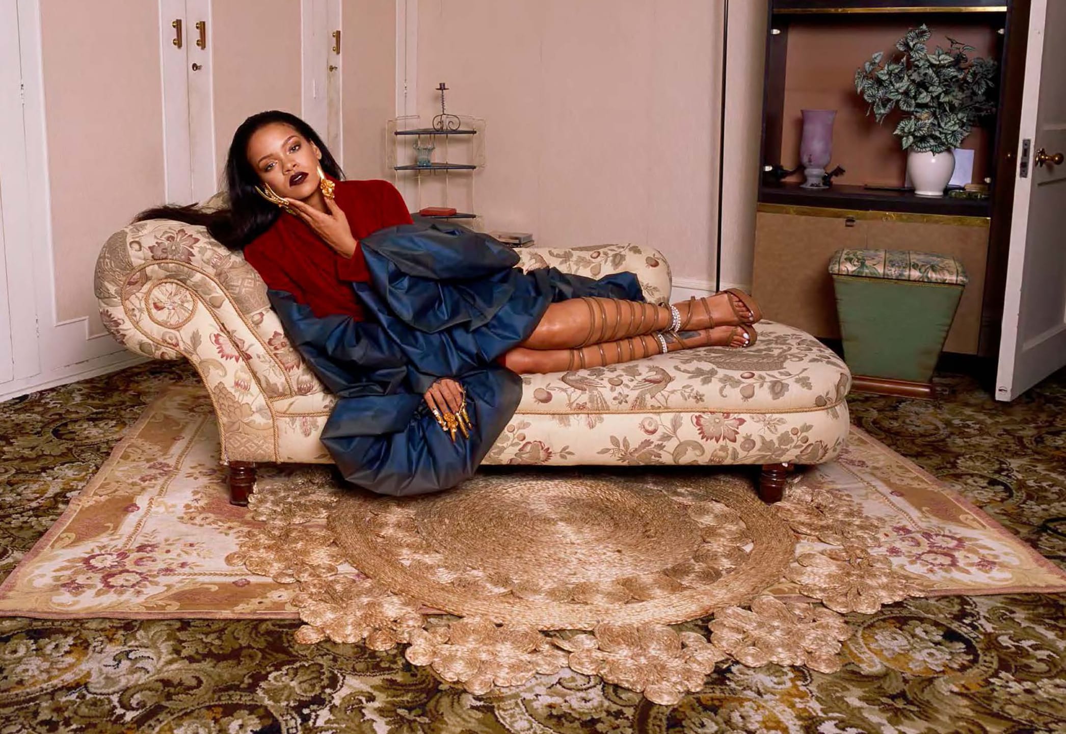 Person lounging on a patterned chaise in a vintage room with ornate carpet and floral decor elements.