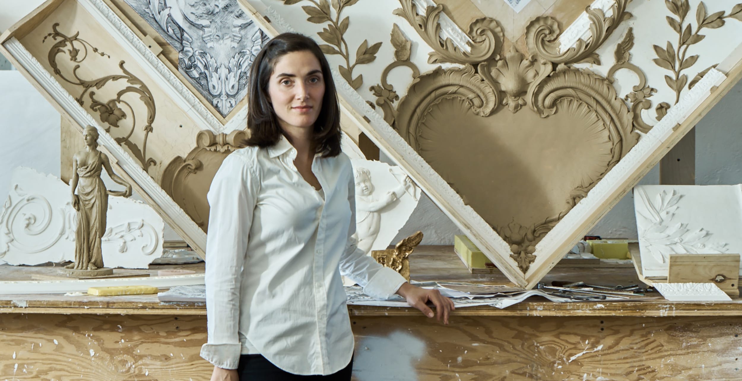 A woman standing beside ornate plasterwork designs in a studio, showcasing detailed craftsmanship and artistry.