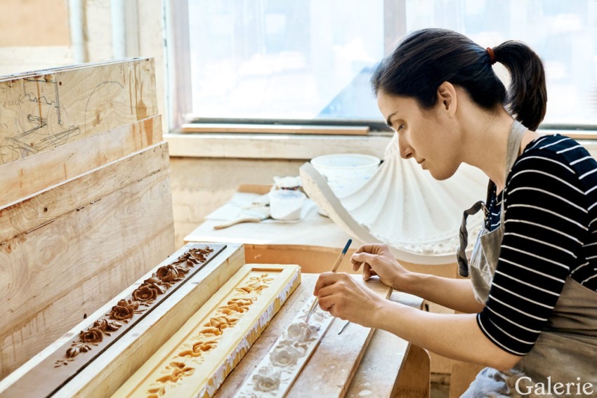 Woman crafting decorative moldings at a workbench in a sunlit workshop, focused on detailing intricate designs.