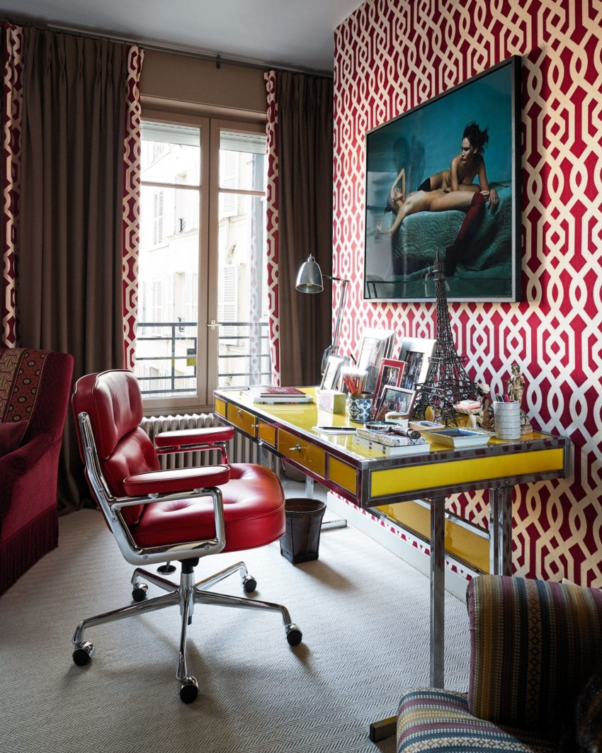 Colorful home office with red patterned wallpaper, yellow desk, red chair, modern artwork, and natural light from large windows.