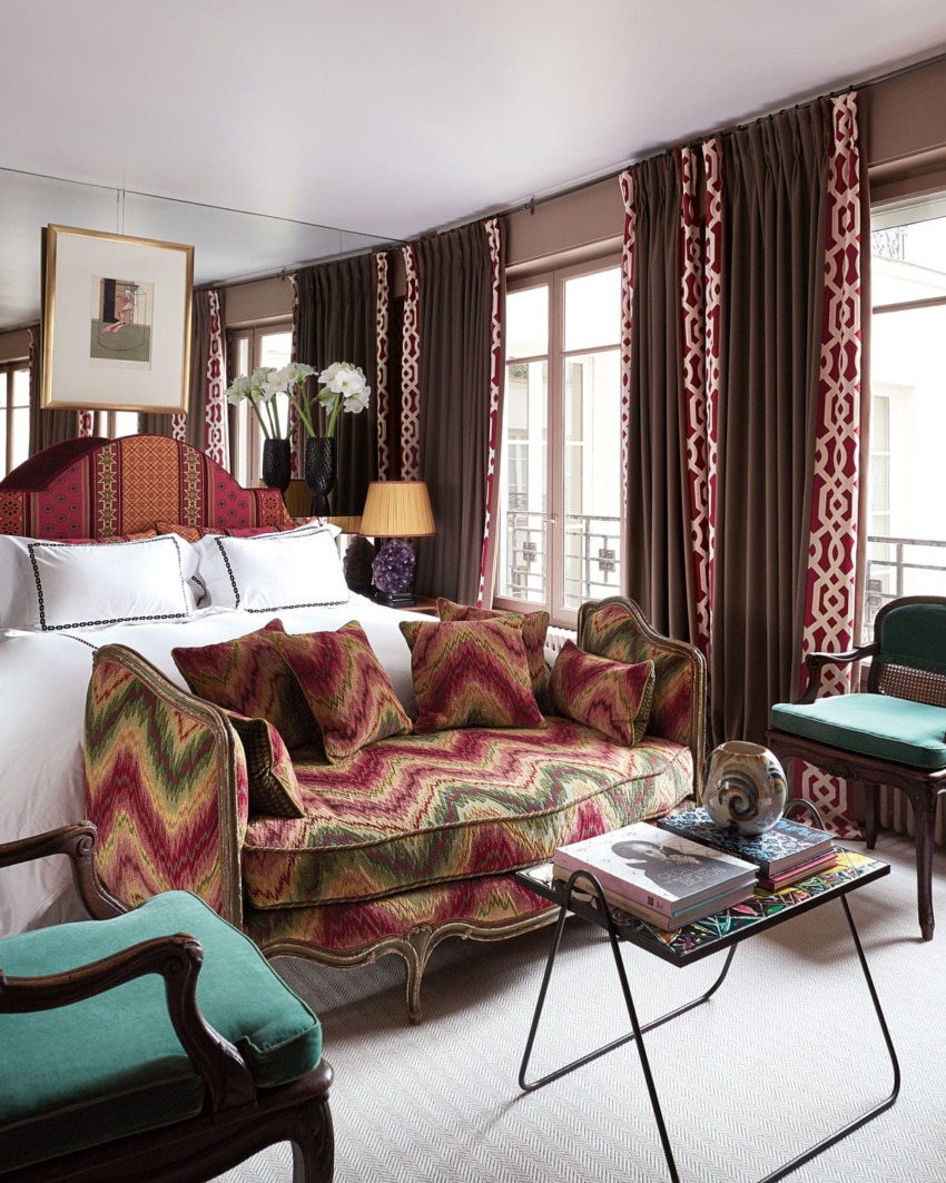 Elegant hotel room with colorful patterned sofa, armchairs, and large windows framed by intricate curtains.