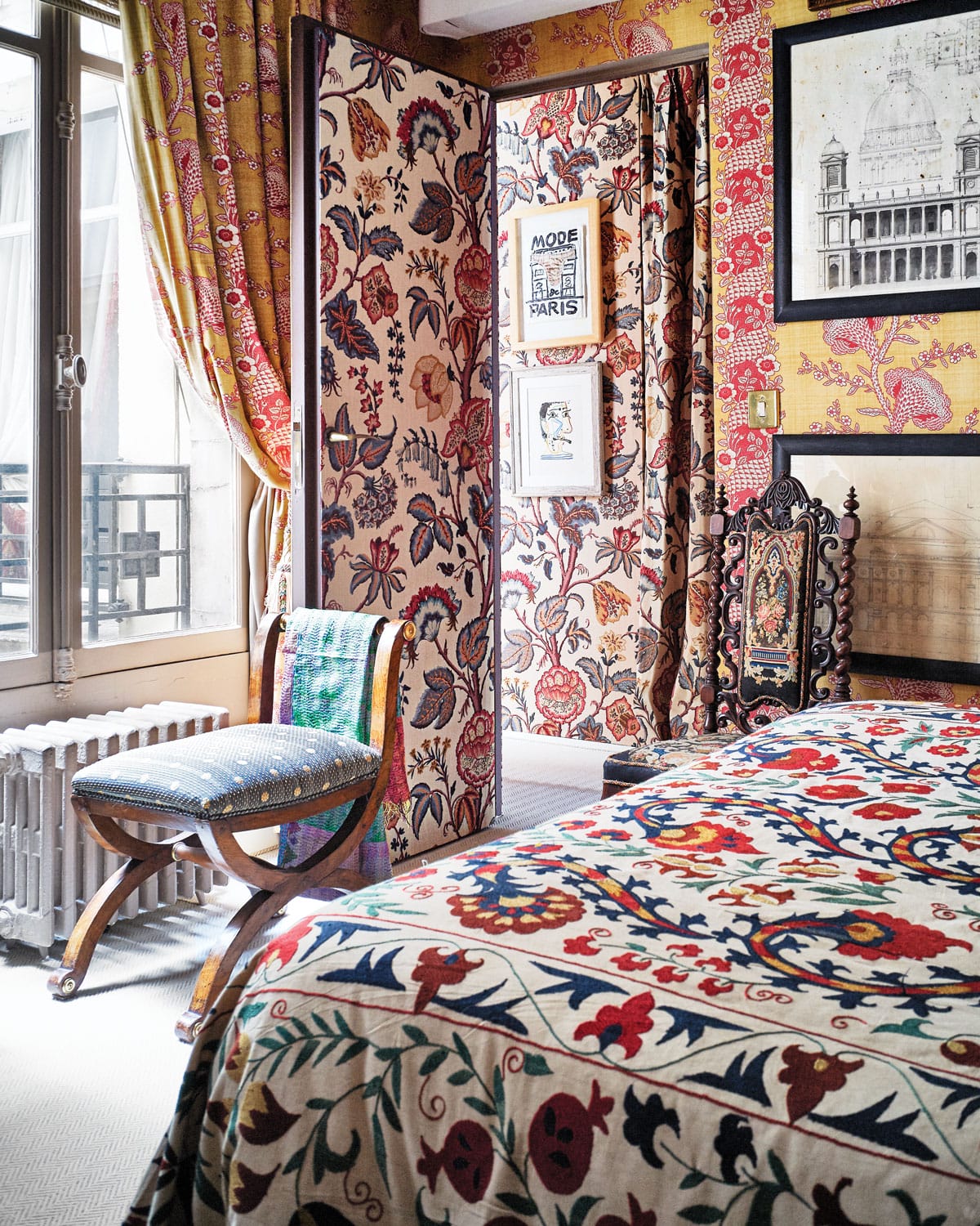 Colorful, eclectic bedroom with patterned textiles, vintage decor, large window, and ornate wooden chair.