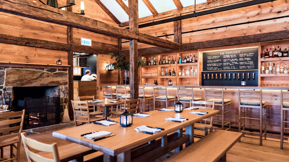 Rustic restaurant interior with wooden beams, cozy fireplace, and a bar area with a chalkboard menu and wooden tables.