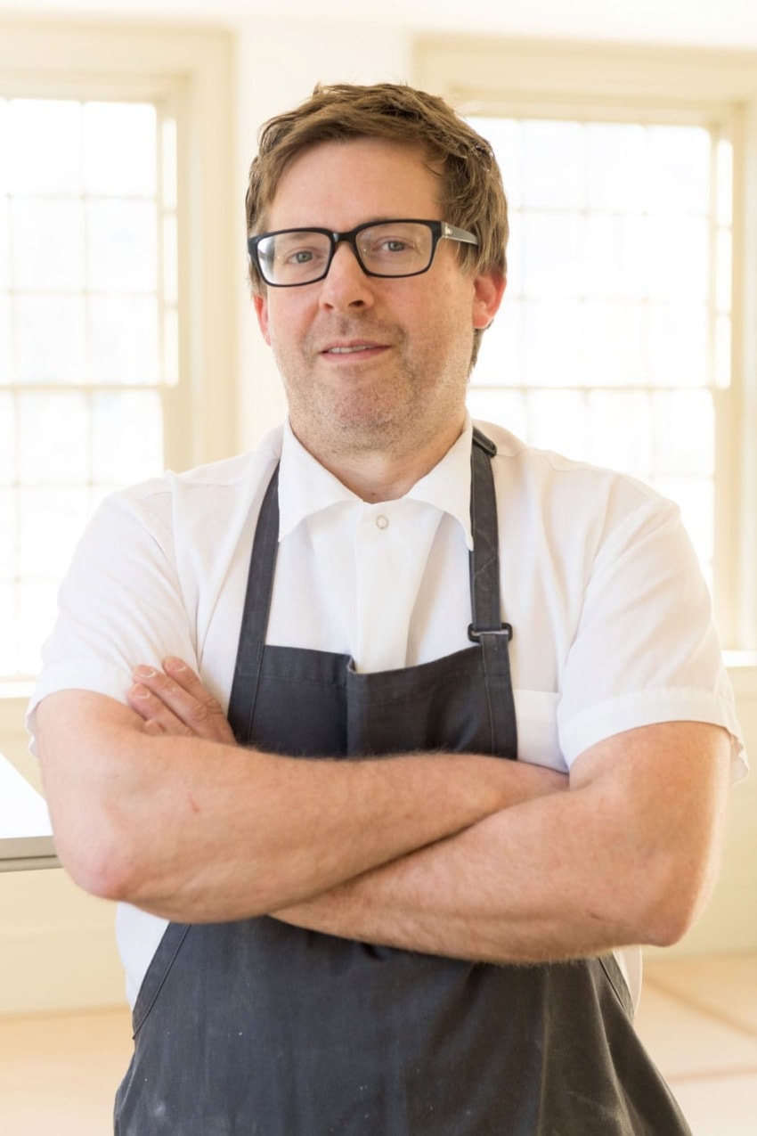 Man in glasses and apron standing with arms crossed in a bright room with large windows.