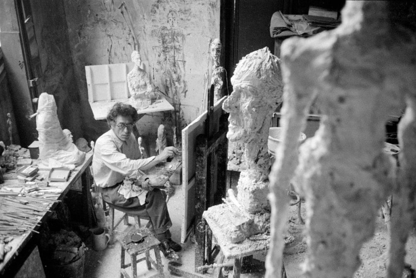 Artist working in a cluttered studio surrounded by large, abstract sculptures and art materials.