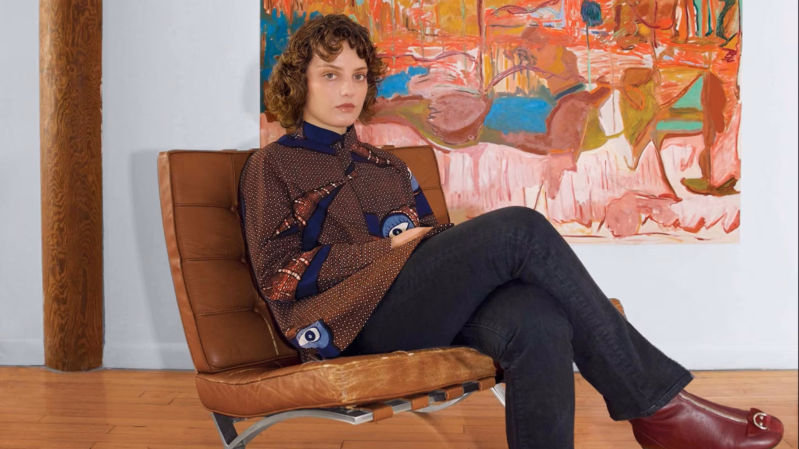 Person sitting on a brown chair with legs crossed, colorful abstract painting in the background