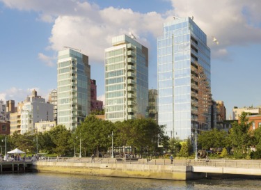 Three modern glass skyscrapers along a waterfront with trees and a clear blue sky in the background.