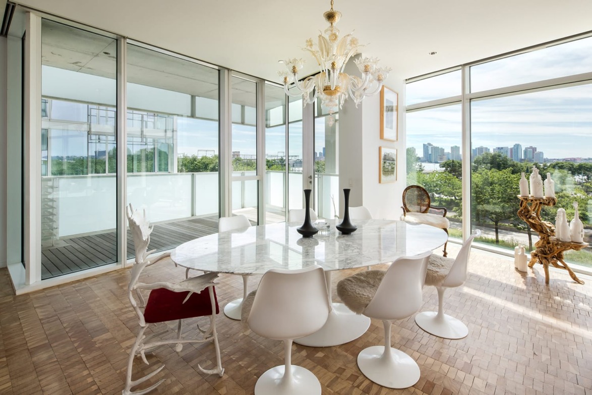 Modern dining room with a marble table, white chairs, and a large chandelier overlooking a cityscape through glass windows.