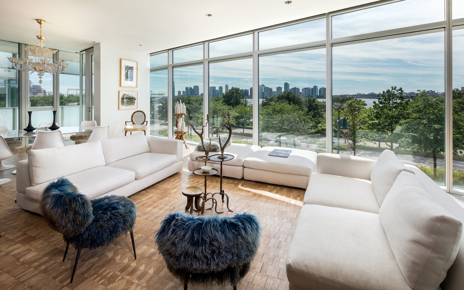 Modern living room with white sofas, blue accent chairs, and city skyline view through large glass windows.