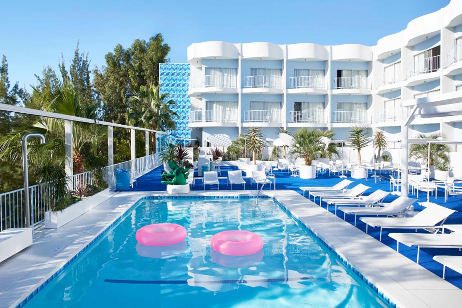 Outdoor hotel pool with pink floaties, surrounded by white lounge chairs and modern building in the background