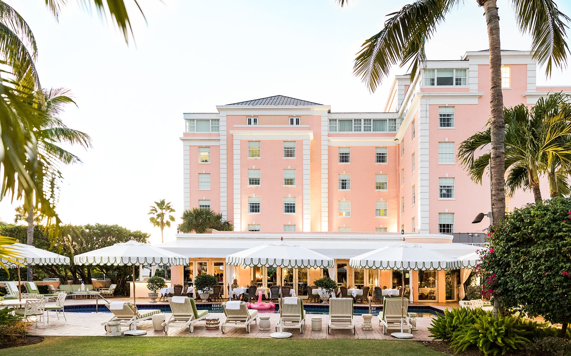 Luxury hotel exterior with palm trees, pool, and white canopied lounge chairs on a sunny day.
