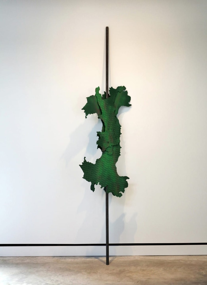 Green abstract metal sculpture mounted on a tall rod against a white wall in a modern art gallery setting.