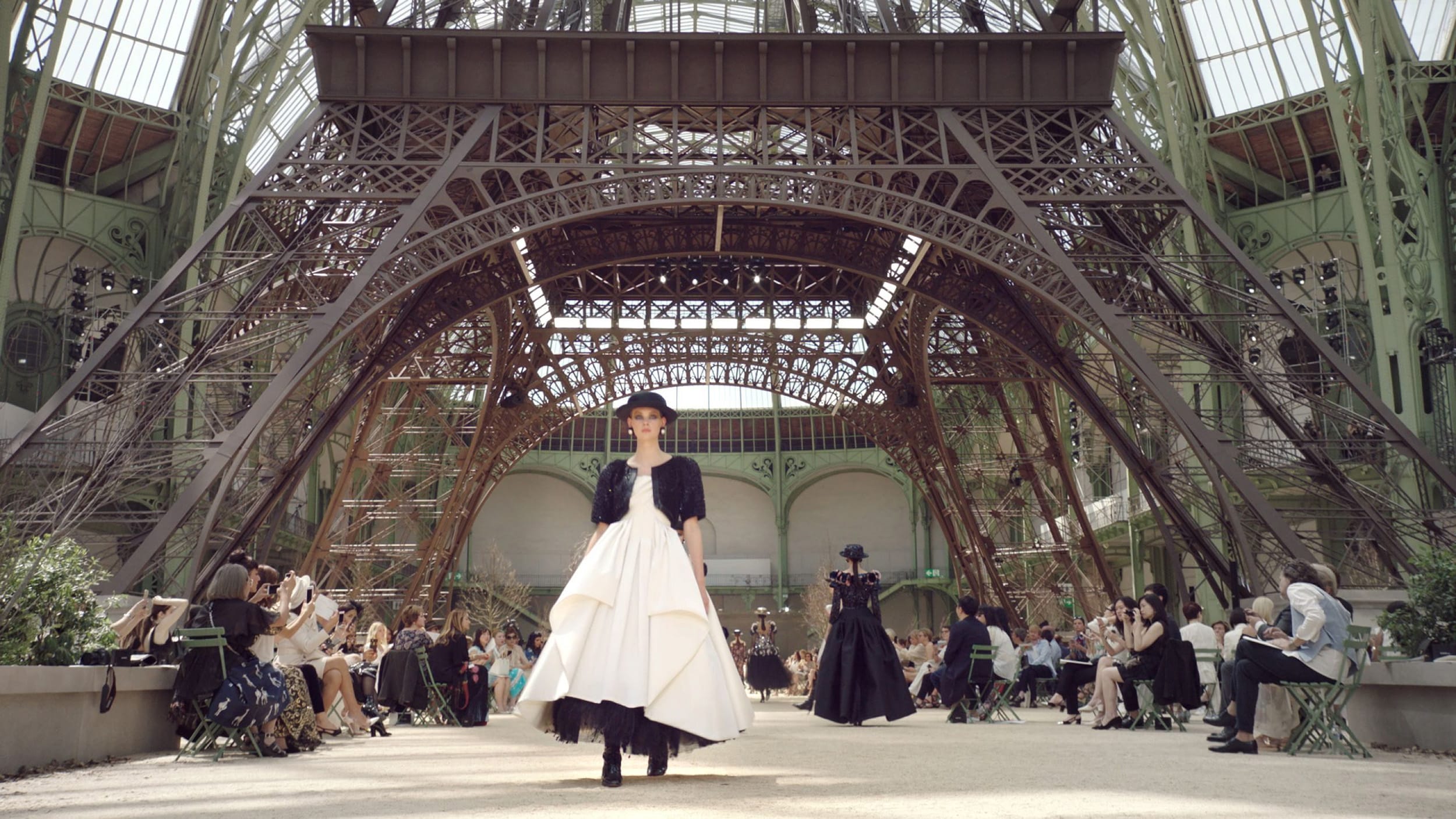 Fashion model walking on a runway under an intricate iron structure during a fashion show, with audience seated nearby.