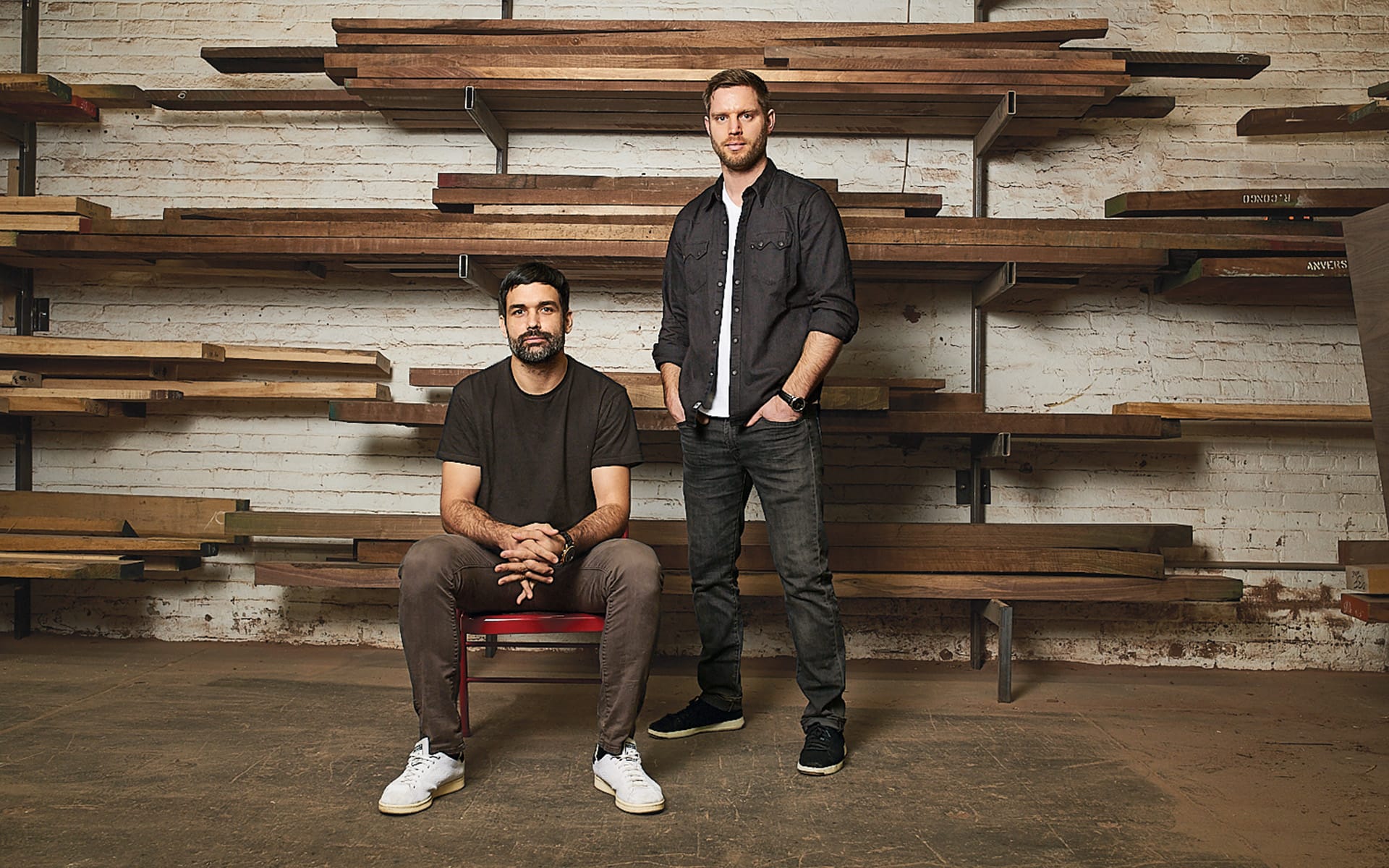 Two men in a workshop with wooden shelves in the background, one seated and one standing, wearing casual clothing.