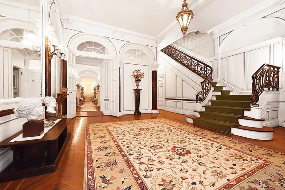 Elegant foyer with ornate staircase, decorative rug, and antique sculptures, featuring intricate woodwork and classic lighting.