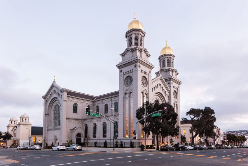 Large church with two golden domes and a clock on a corner street, surrounded by cars and trees, on a cloudy day.