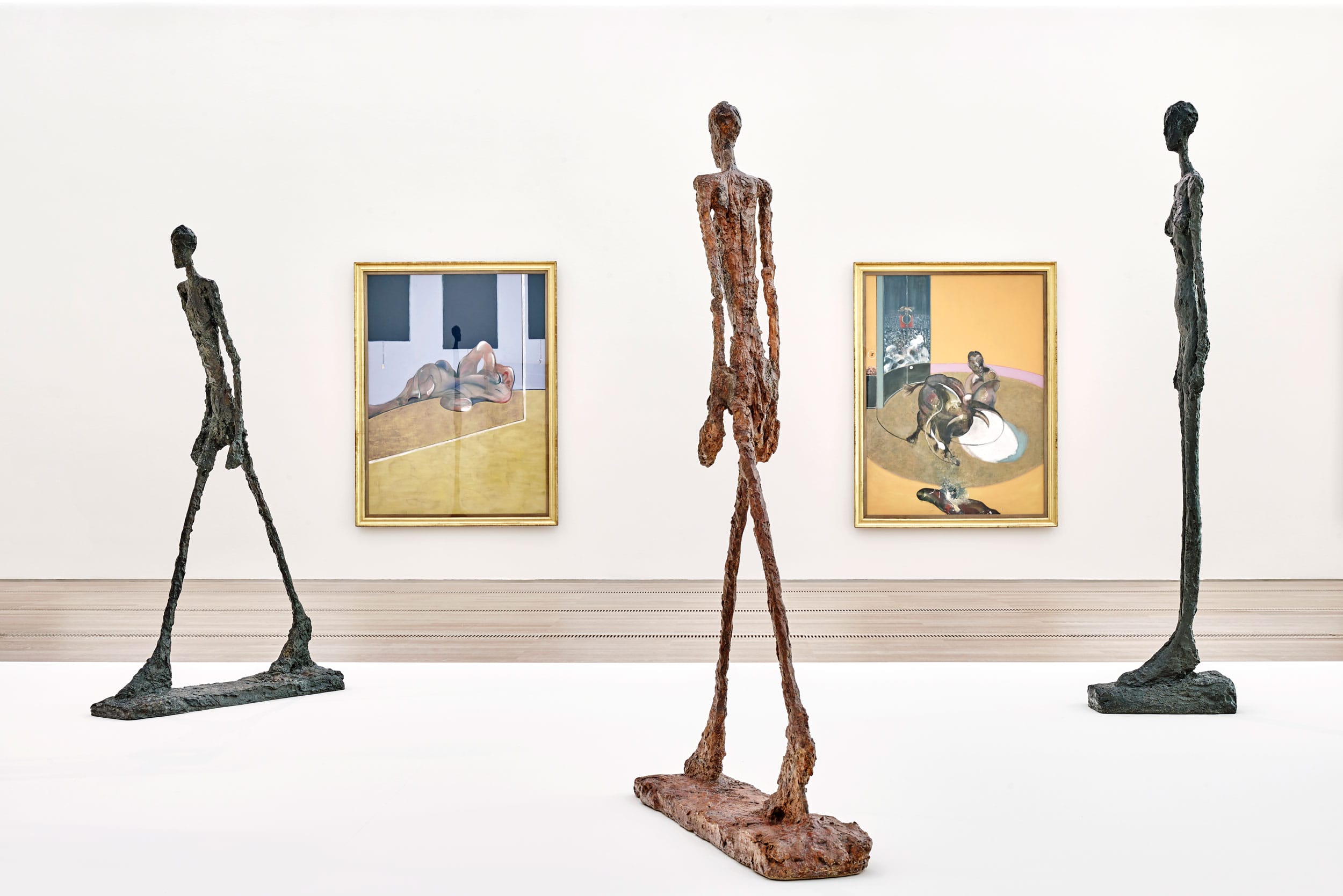 Sculptures of elongated figures in a gallery with paintings hanging on the wall in the background.