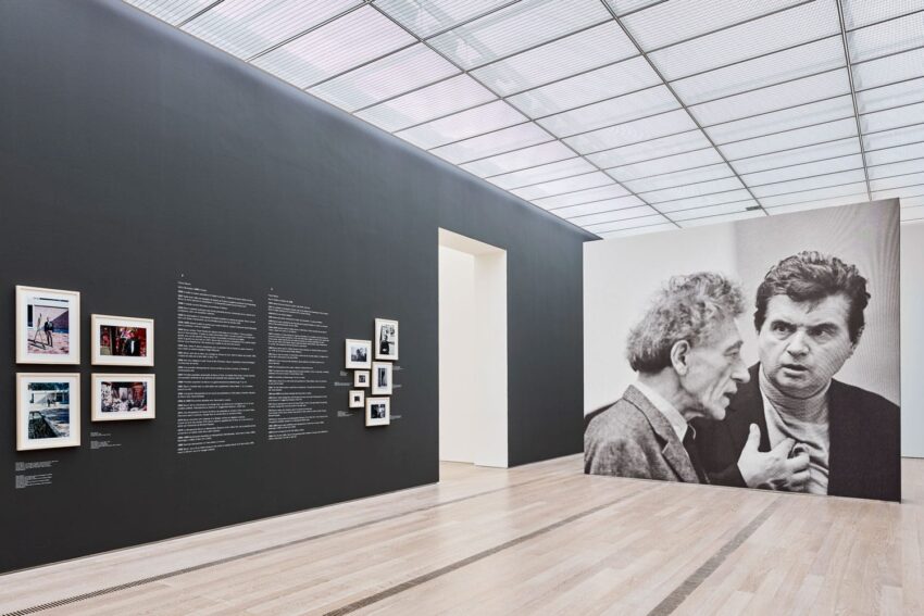 Art gallery interior with photographs on walls, a large black-and-white portrait, and natural light from the ceiling.