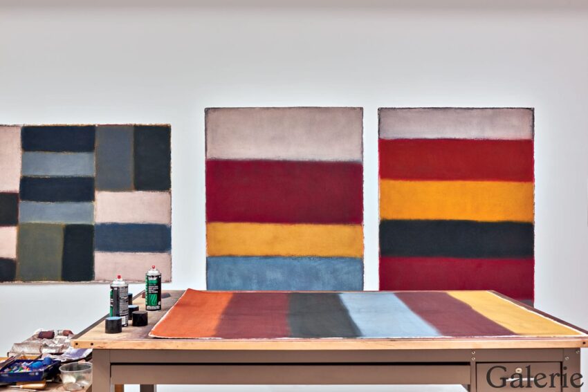 Abstract paintings with colorful geometric patterns on a gallery wall, art supplies on a table in front.