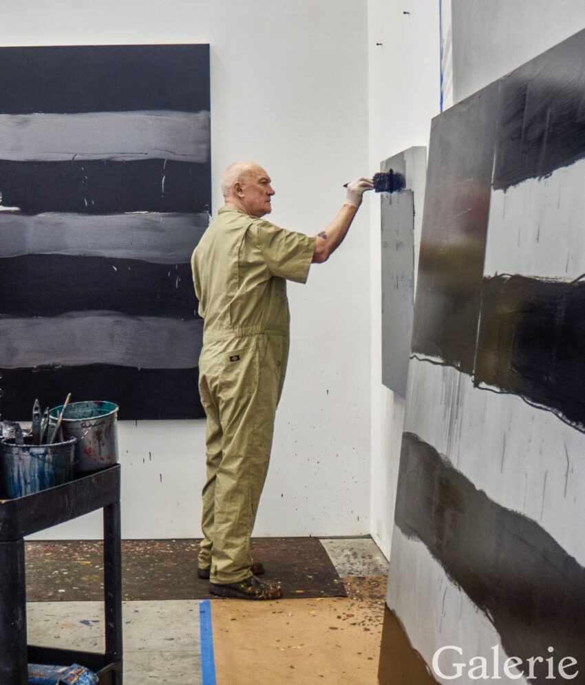 Artist painting abstract artwork in a studio, surrounded by large canvases with bold black and gray stripes.