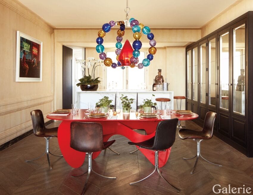 Dining room with a vibrant red table, colorful chandelier, modern chairs, and elegant decor elements for a stylish setting.