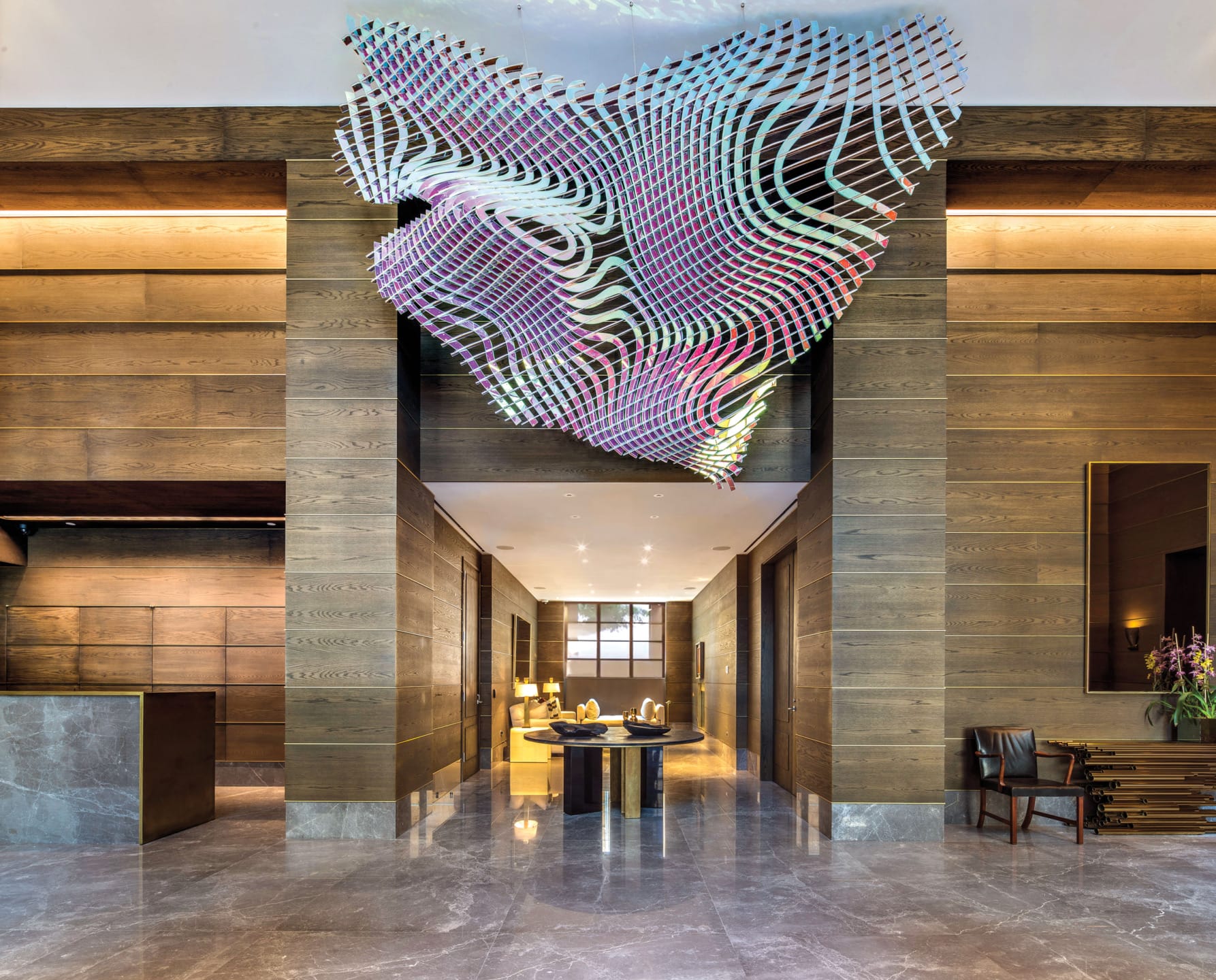 Modern hotel lobby with artistic ceiling installation, wooden walls, marble floors, and seating area.