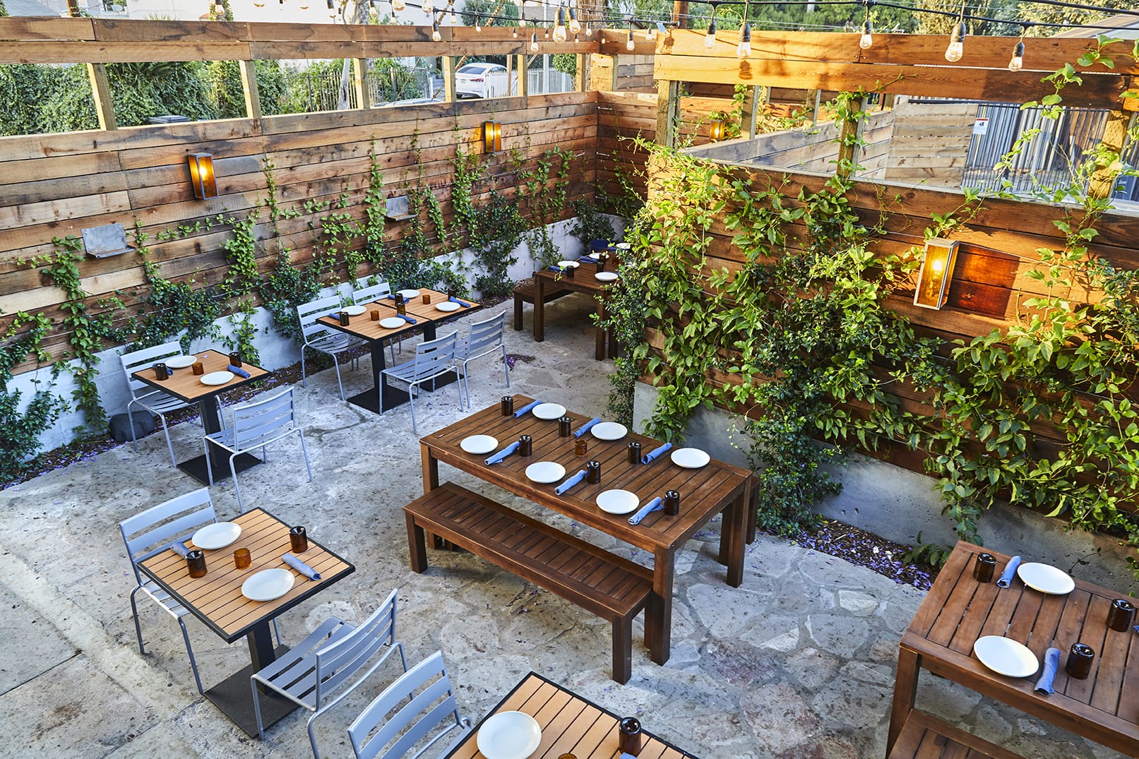 Outdoor restaurant patio with wooden tables and chairs, surrounded by greenery and hanging lights for ambiance.
