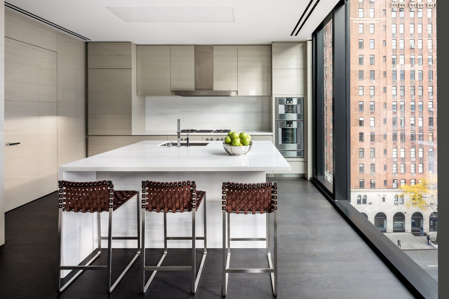 Modern kitchen with large island, woven stools, stainless steel appliances, and floor-to-ceiling windows overlooking cityscape.
