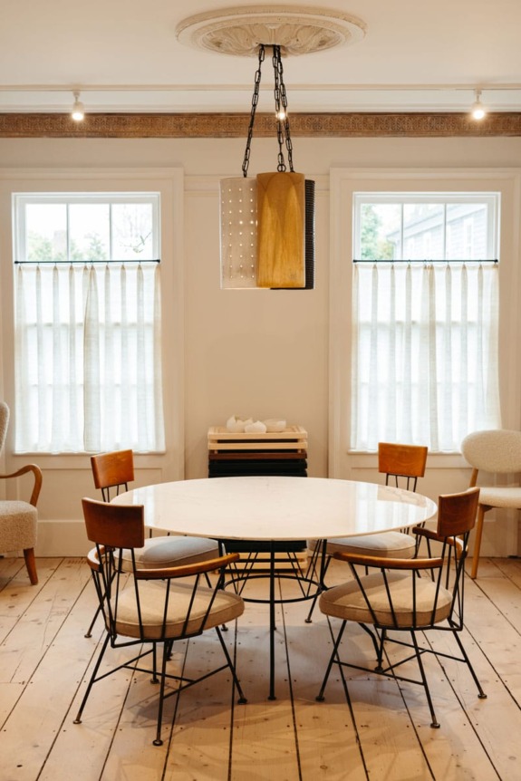 Modern dining room with round table, four wooden chairs, large windows, sheer curtains, and a geometric pendant light.