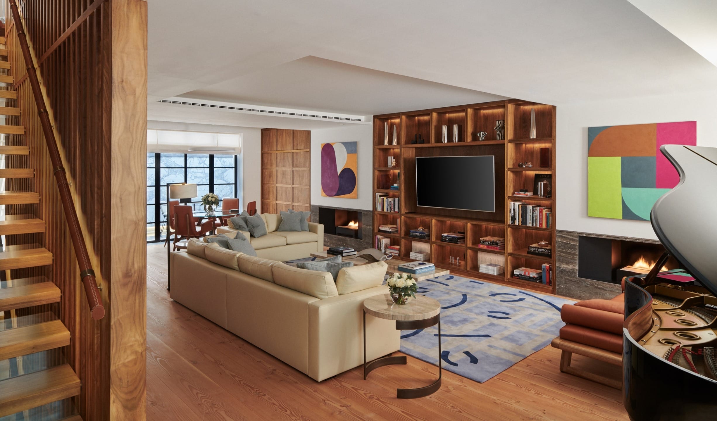 Modern living room with a beige sectional sofa, large TV, abstract art, bookshelves, piano, and a wooden staircase.