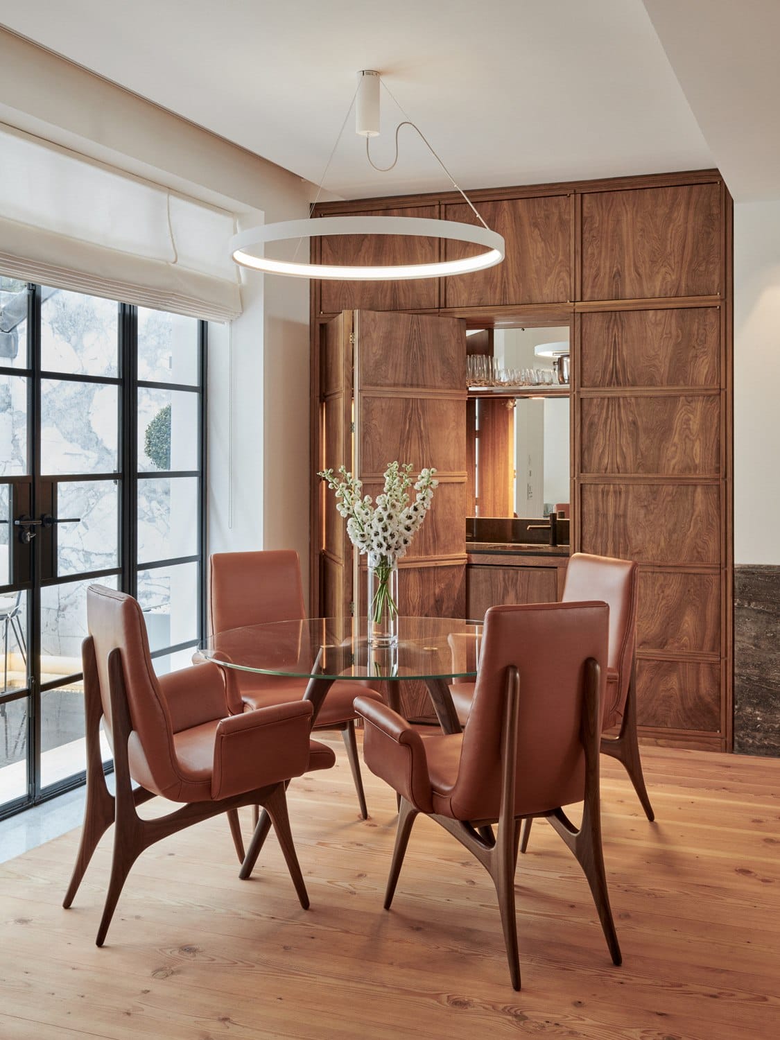 Modern dining room with leather chairs, glass table, circular light fixture, and wooden paneling accent wall.