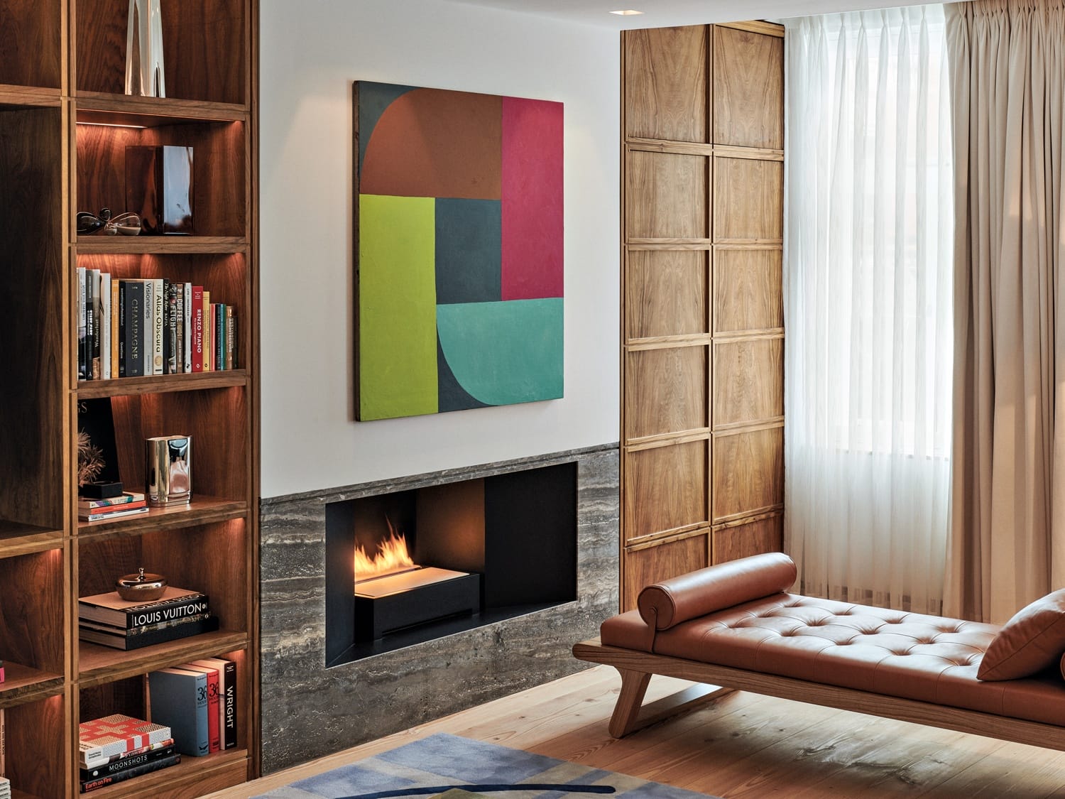Modern living room with a colorful abstract painting, built-in wooden bookshelves, a lit fireplace, and a leather chaise lounge.