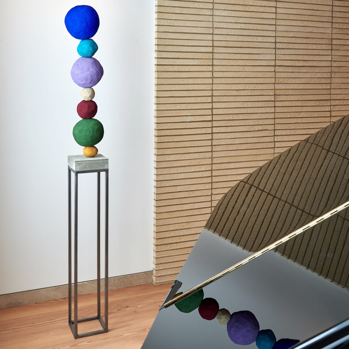 Colorful stacked spheres sculpture on tall metal stand beside reflective piano in a room with wooden floor and panel walls.