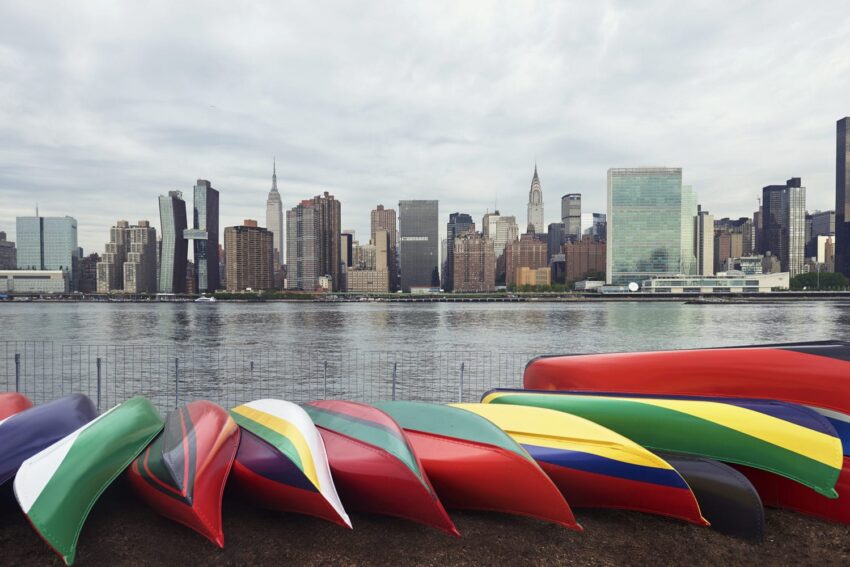 Colorful canoes on shore with New York City skyline in the background under a cloudy sky.
