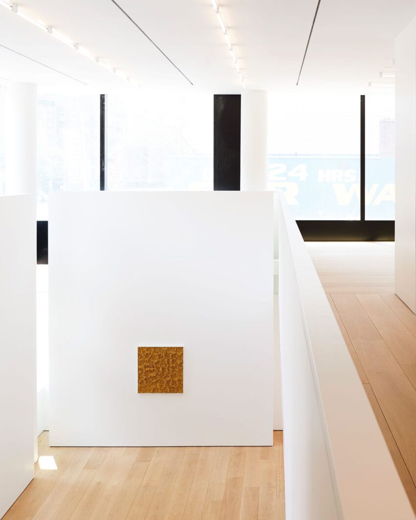 Minimalist art gallery interior with wooden floors, large windows, and a small gold square artwork on a white wall.