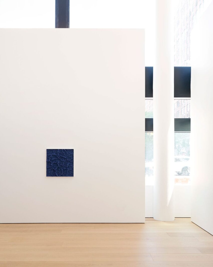 Minimalist gallery interior with a small dark blue textured artwork on a large white wall, wooden floor, and natural light.