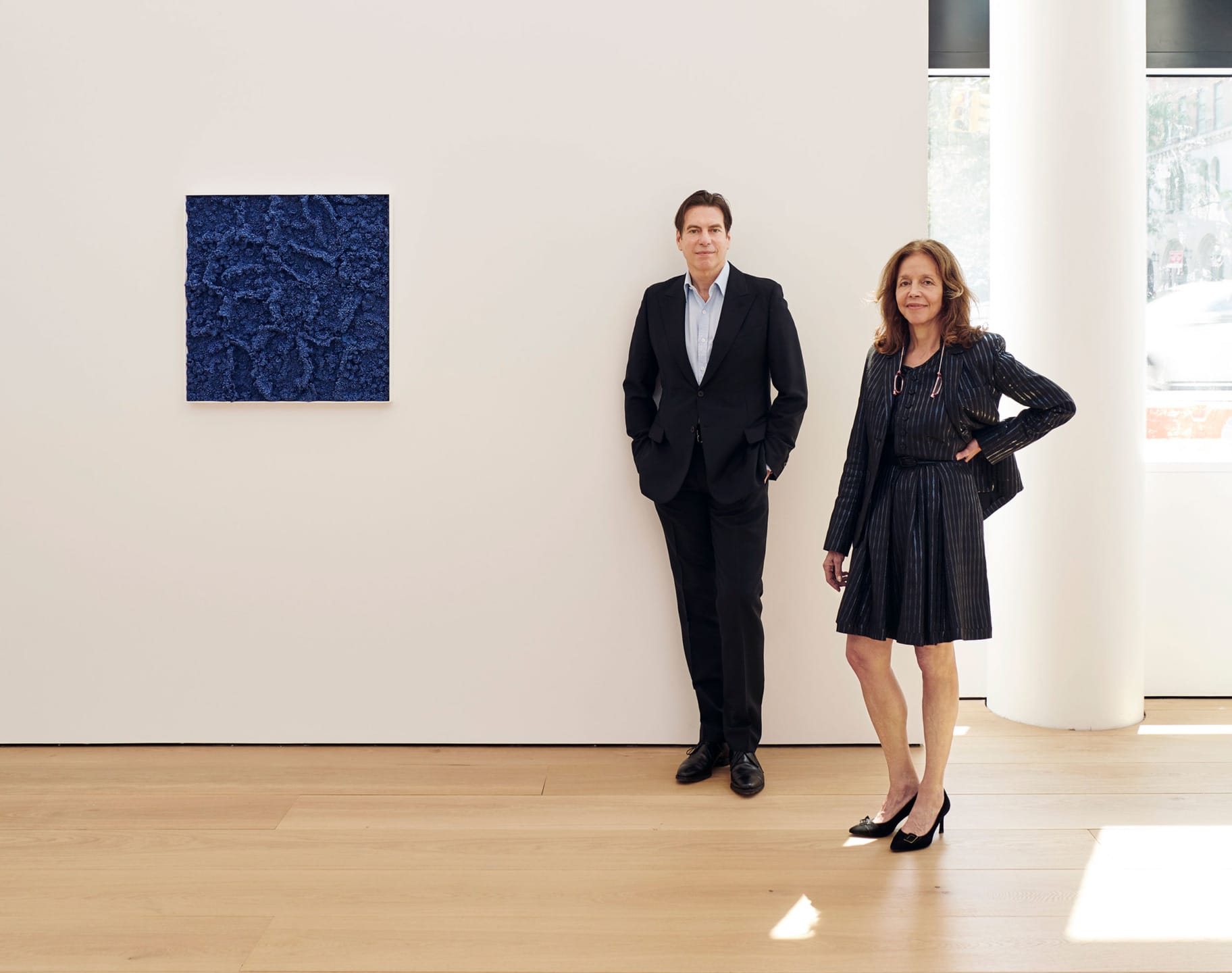 Two people in formal attire standing near a blue abstract painting on a gallery wall with wooden flooring.
