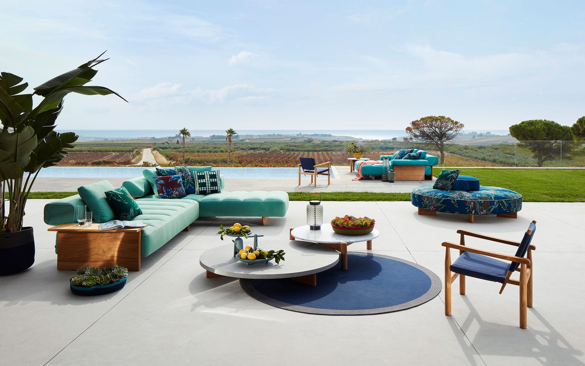 Modern outdoor patio with turquoise furniture, round coffee tables, and scenic view of landscape with pool and distant horizon.